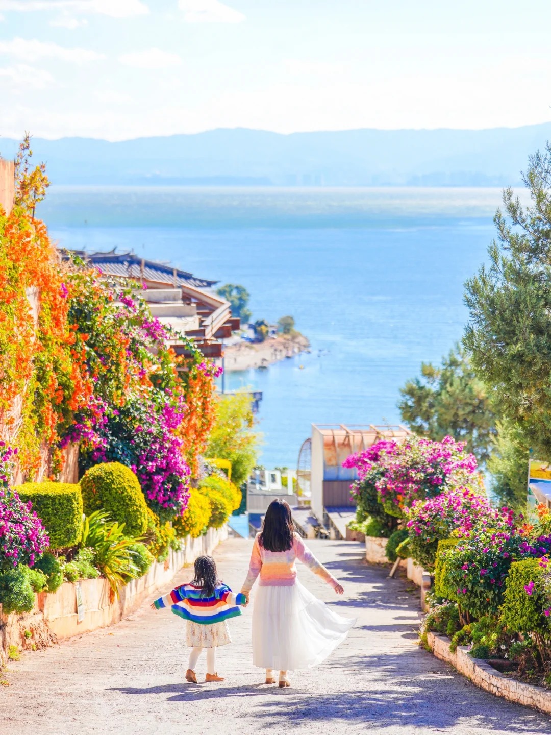 第 1 秒:画面锁定原图,母女牵手沿花路向前走,镜头同步向前轻移,两侧繁花细节渐清晰。第 2 秒:母女裙摆随步伐轻扬,镜头微微抬升,远处湖景开始显露。第 3 秒:镜头持续向前跟拍同时缓慢向上拉升,母女手臂舒展的姿态更突出。第 4 秒:镜头继续缓慢向上拉升,视角从平视转为高角度俯瞰。第 5 秒:视角从高角度俯瞰,花路、母女与湖景构成色彩饱满的远景画面。